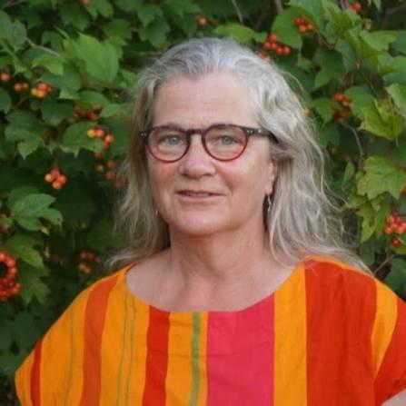 La Dre Alison Eyre sourit et porte des lunettes ainsi qu’un chandail rayé jaune, orange et rose. L’arrière-plan montre un grand arbuste à baies vert, avec des feuilles vertes et des baies rougeâtres et jaunes.