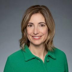 Dre Claire Kendall sourit à la caméra, vêtue d’un haut vert vif à col, devant un arrière-plan gris.
