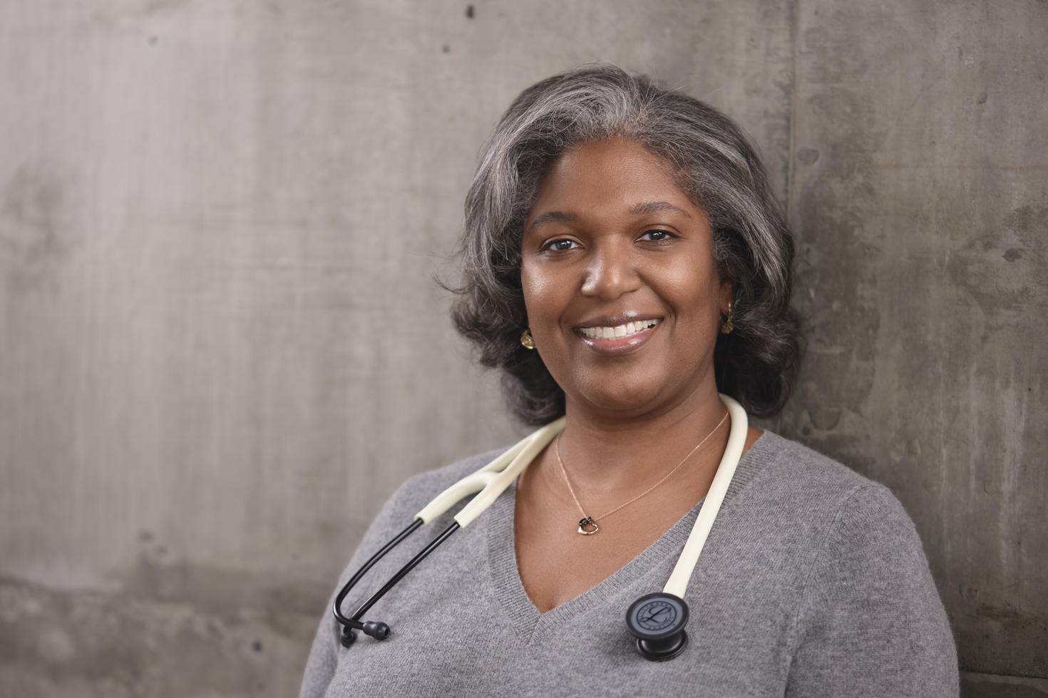 Portrait professionnel du Dre Denice Lewis, souriante, aux cheveux poivre et sel tombant jusqu'aux épaules, vêtue d'un pull gris à col en V et portant un stéthoscope, posant devant un mur en béton.