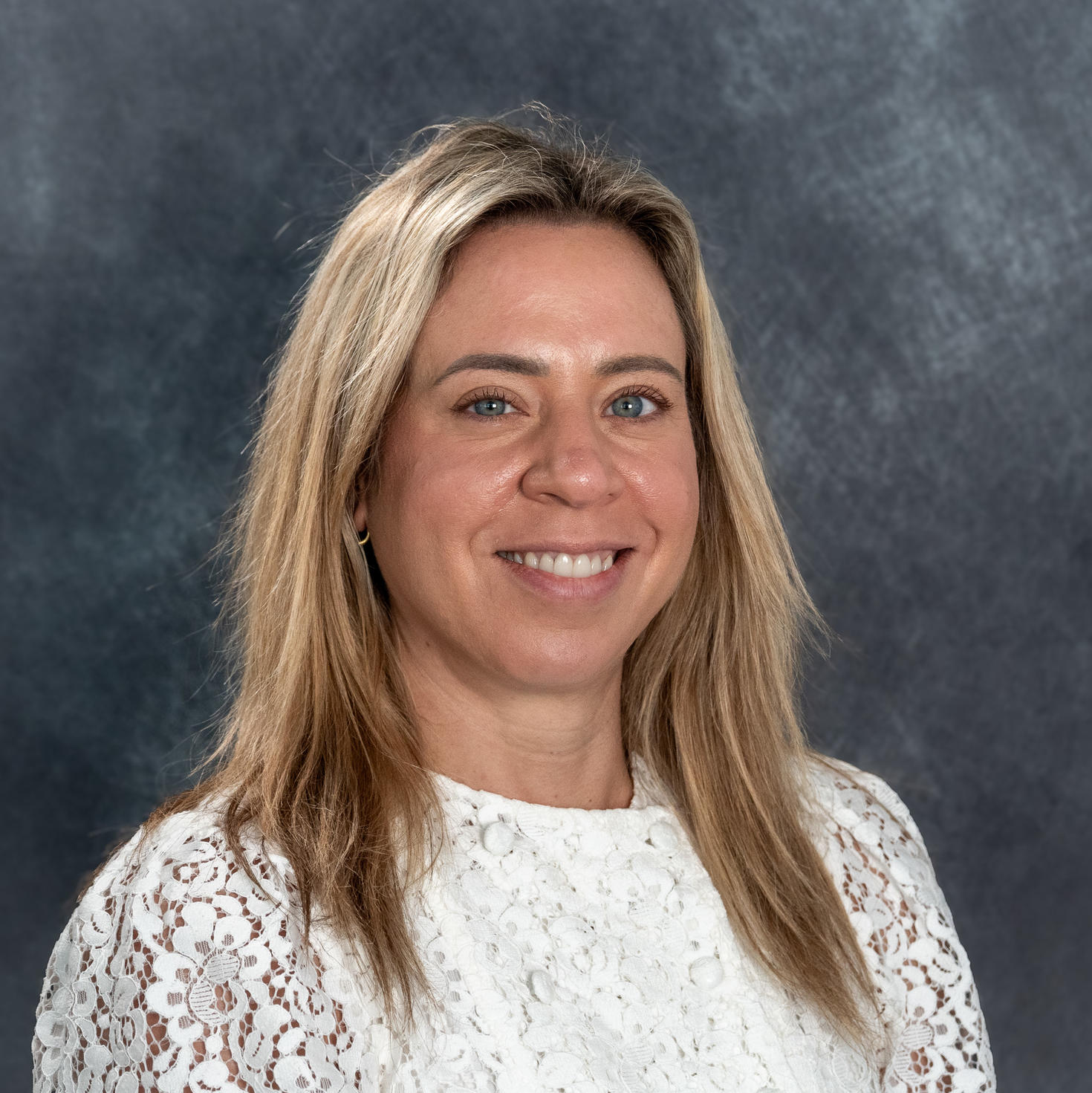 Marisa Duval, gestionnaire principale de projet, sourit et porte un haut blanc en dentelle, posant devant un arrière-plan bleu marine à motifs.