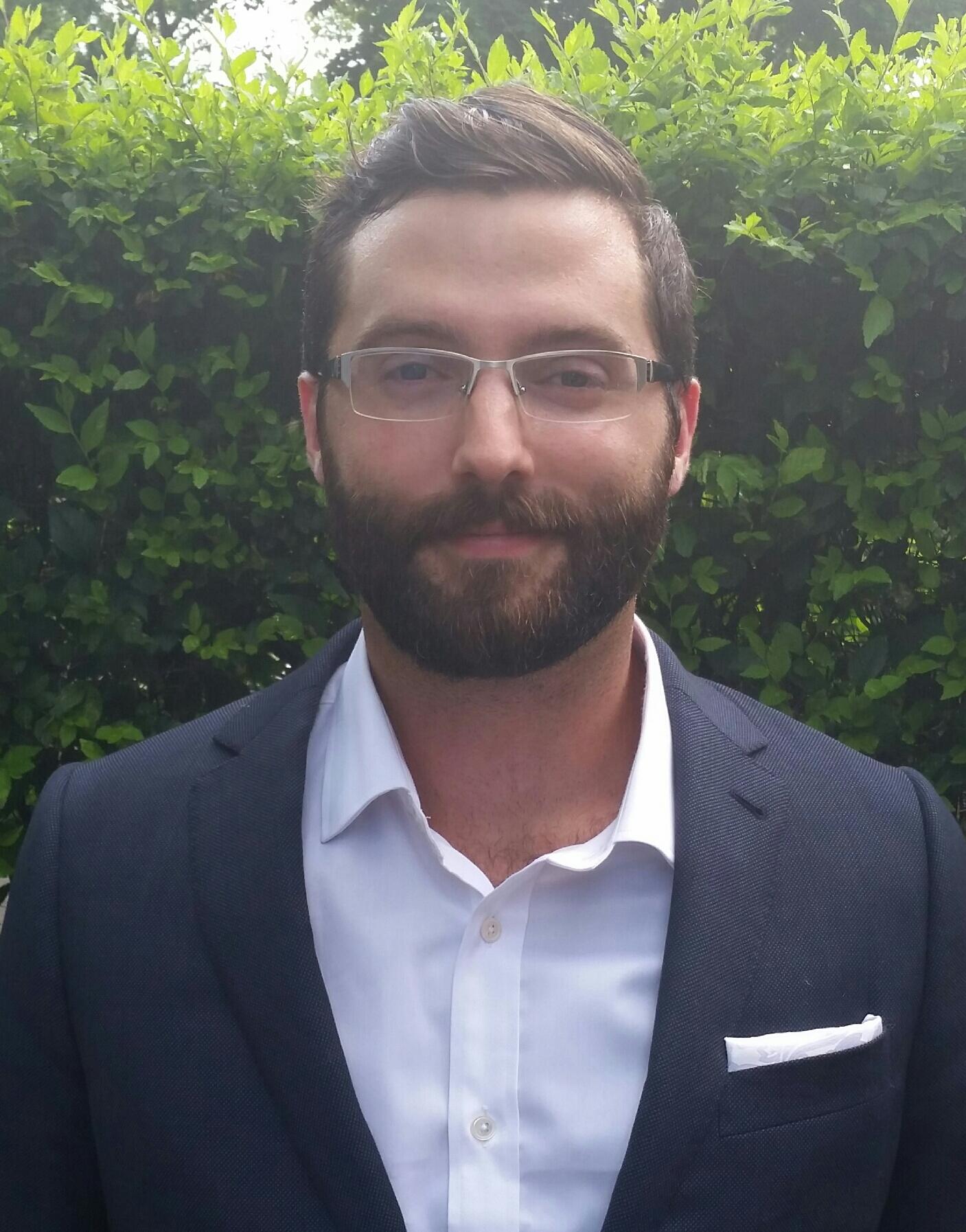 Zack Van Allen, vêtu d'un costume noir et d'une chemise blanche à col, devant un fond de verdure