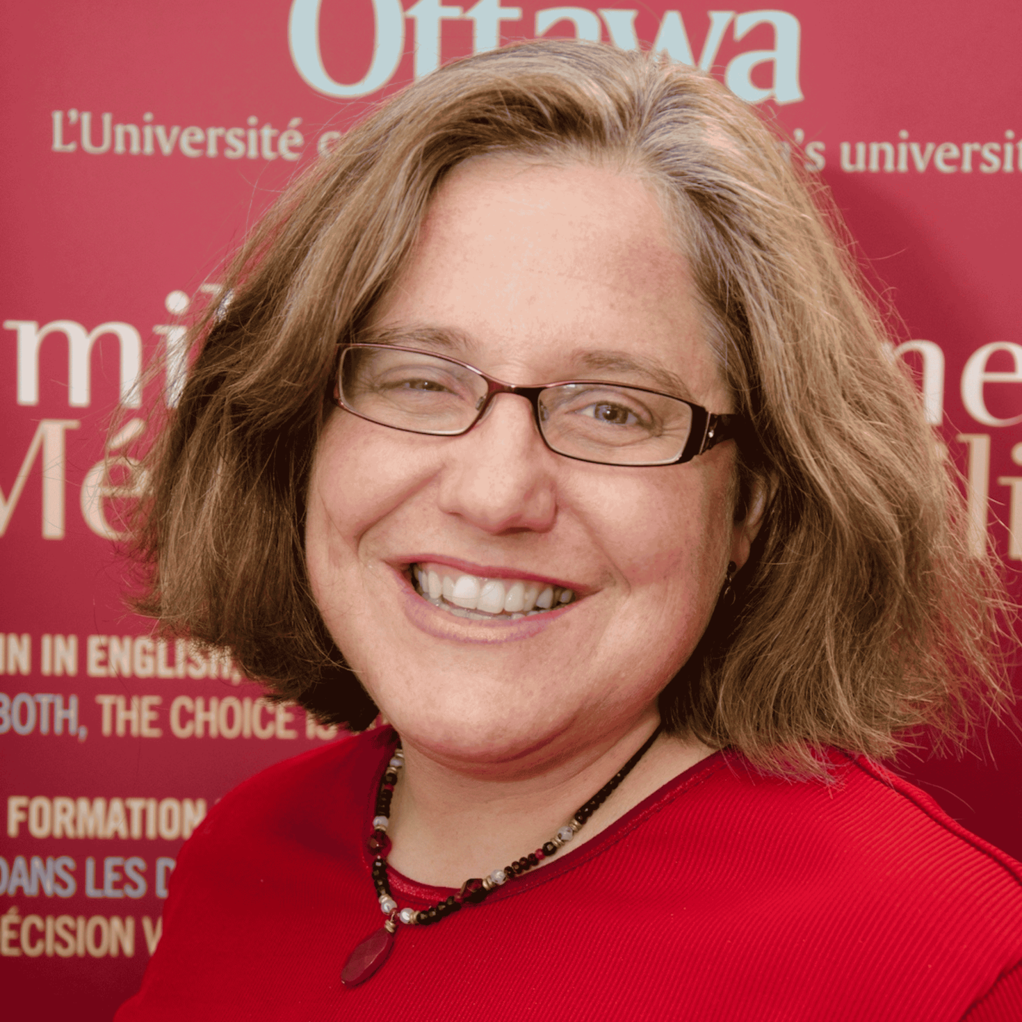 La Dre Carol Geller sourit et porte un haut rouge vif ainsi que des lunettes. Elle pose devant un arrière-plan rouge de l’Université d’Ottawa, orné de diverses inscriptions blanches.