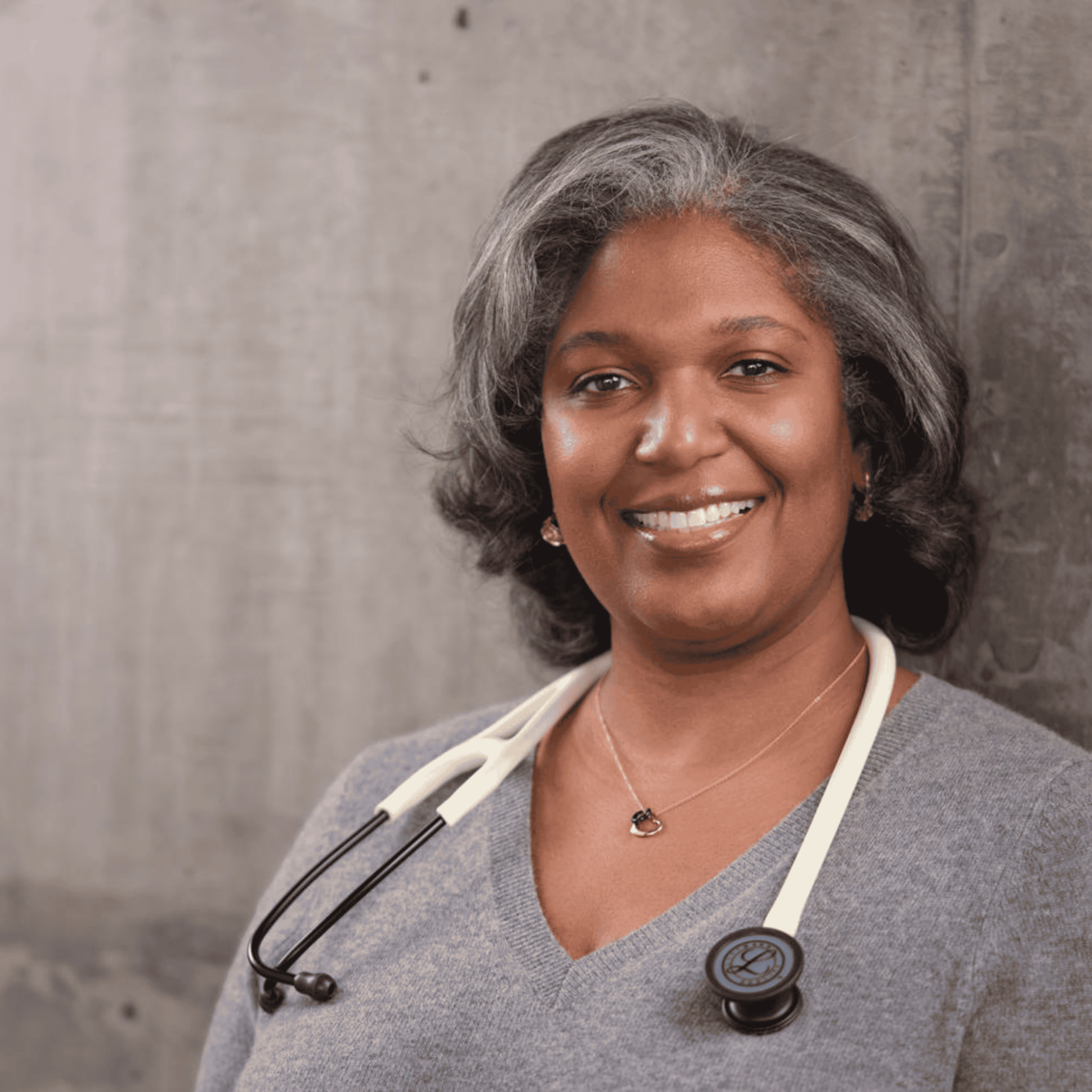 Portrait professionnel du Dre Denice Lewis, souriante, aux cheveux poivre et sel tombant jusqu'aux épaules, vêtue d'un pull gris à col en V et portant un stéthoscope, posant devant un mur en béton.