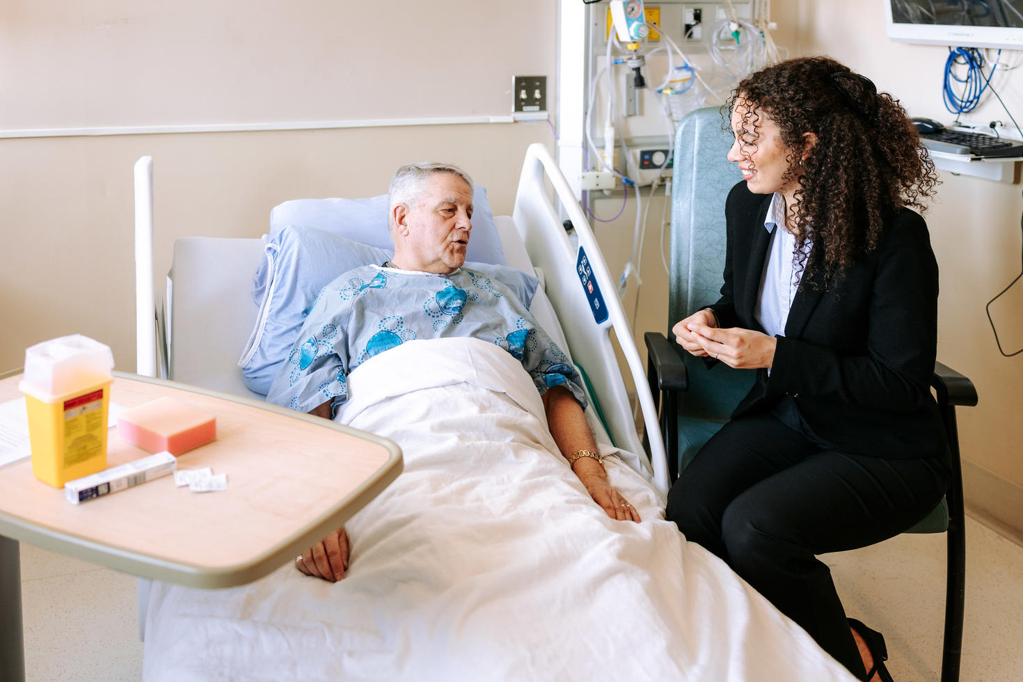 Une pharmacienne vêtue d'un blazer foncé sourit en s'entretenant avec une patiente âgée allongée dans un lit d'hôpital, un gobelet à médicaments à la main.