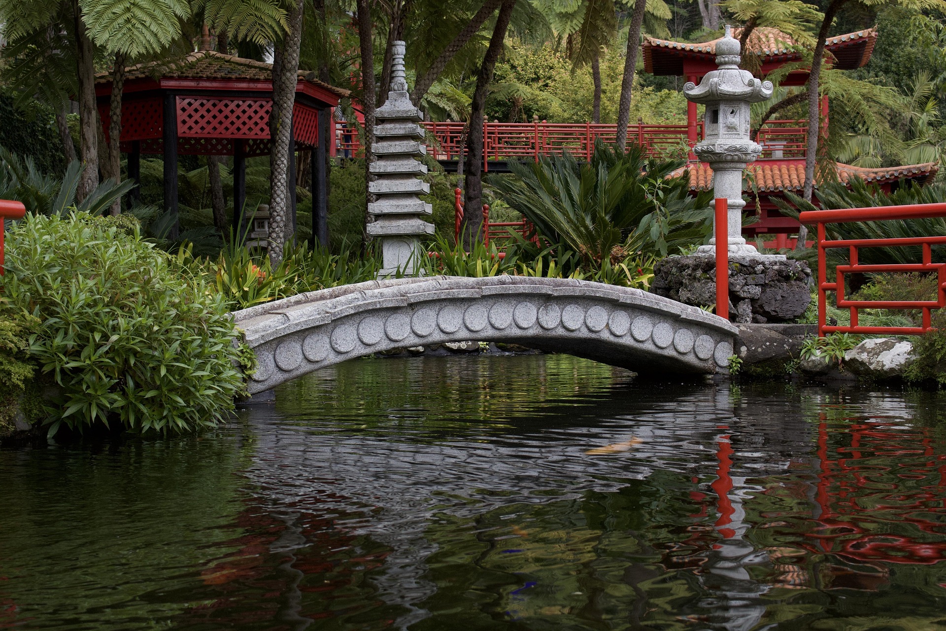 Un pont de pierre en arche enjambe un étang paisible dans un jardin luxuriant; une pagode rouge est en arrière-plan.