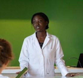 Une femme en blouse blanche debout derrière un comptoir, parlant à une personne assise en face d'elle, avec un mur vert vif en arrière-plan.