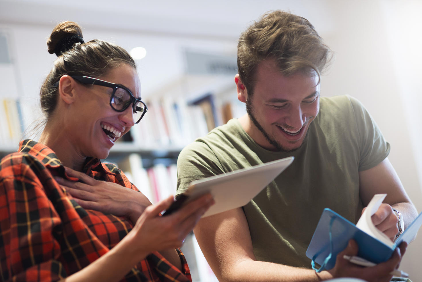 Deux étudiants riant ensemble, un avec un livre à la main, l'autre avec une tabelette.