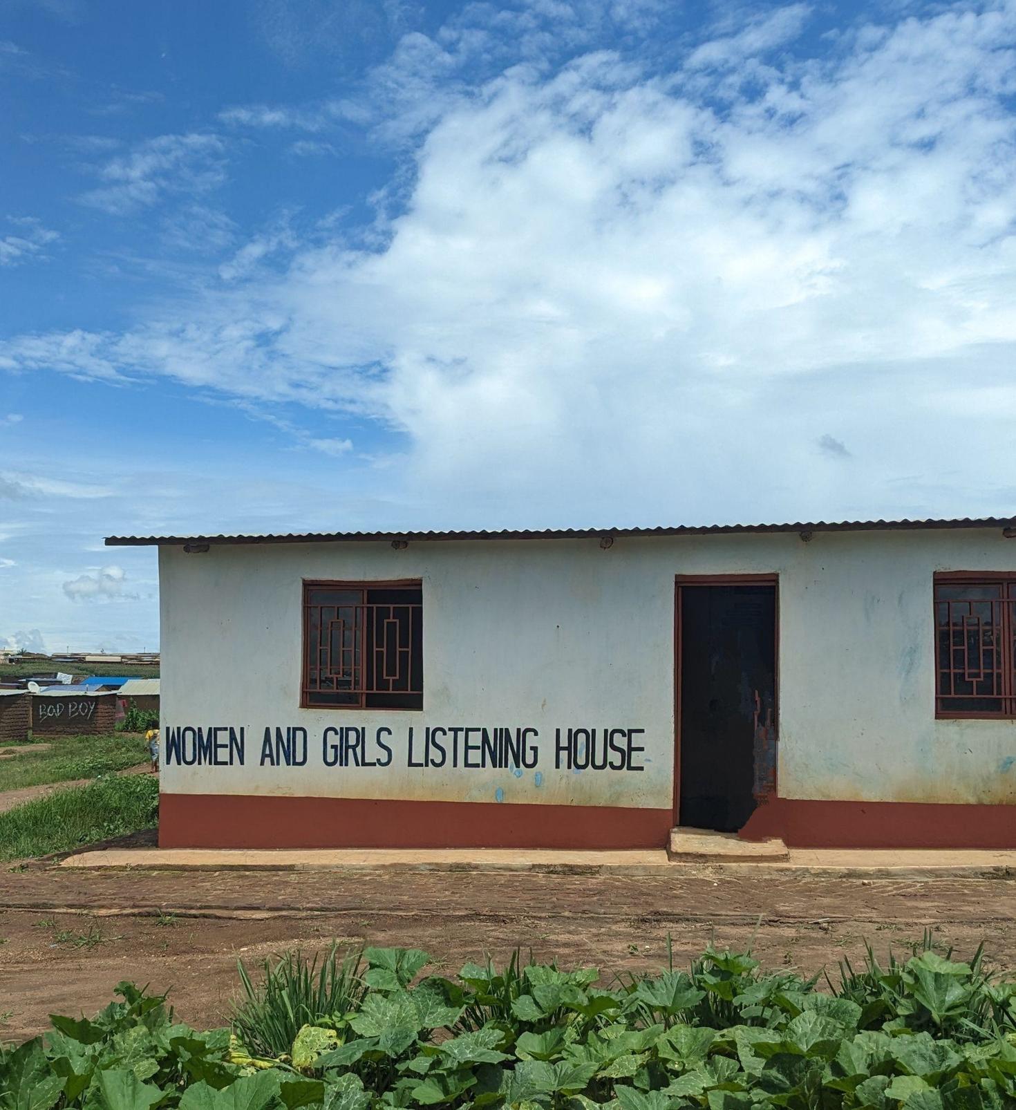 Small building in the middle of a field. White and orange walls with written "Women and girls listening house" on a wall. Blue sky in the background