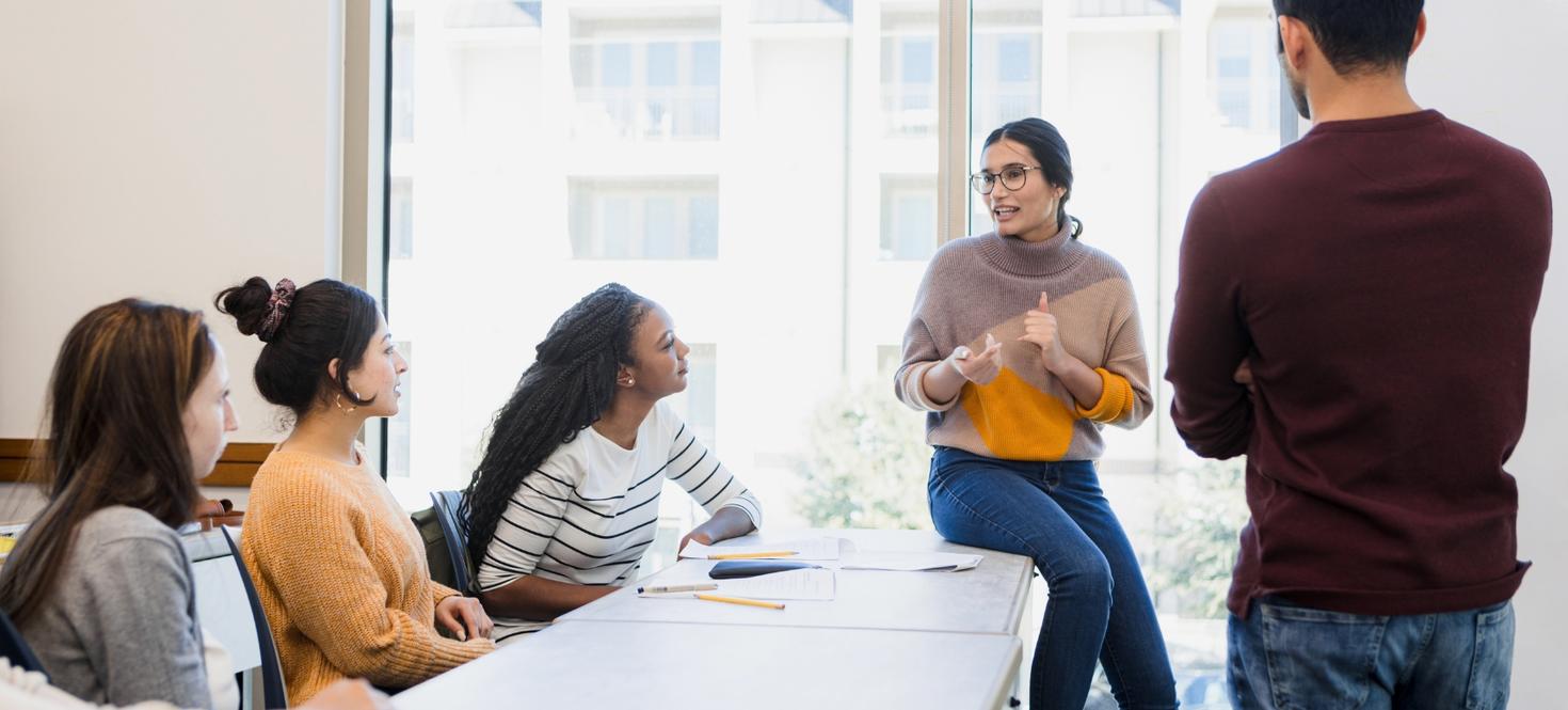 Trois étudiants en criminologie sont assis et un autre se tient debout face à un autre étudiant qui s'adresse à eux dans une salle de classe bien éclairée.