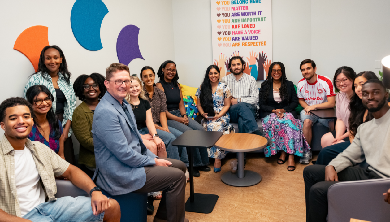 Un groupe d'étudiantes, d'étudiants et de membres du personnel est réuni dans le nouvel espace d'affinité, assis ensemble et souriant vers l'objectif. La pièce comprend des sièges confortables, une décoration colorée et une affiche mettant de l'avant les valeurs d'appartenance, de respect et d'inclusion.
