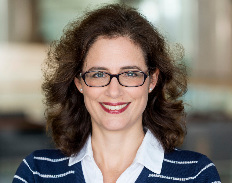 Portrait professionnel d’une femme souriante aux cheveux bruns mi-longs et portant des lunettes, une chemise blanche à col et un chandail bleu marine rayé.