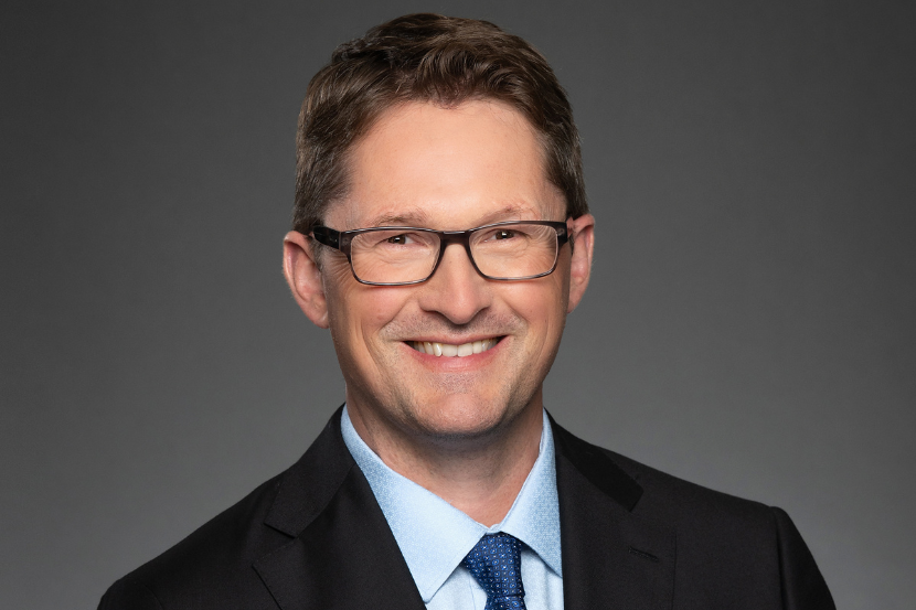 Portrait professionnel d’un homme souriant portant des lunettes, un complet foncé, une chemise bleu pâle et une cravate bleue, sur un fond gris foncé.