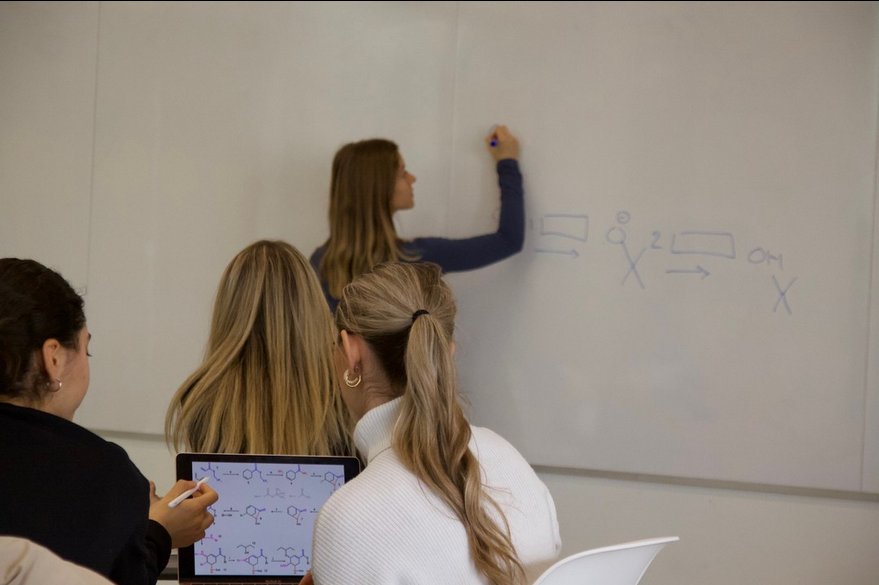 Une étudiante écrit sur un tableau blanc pendant que d'autres étudiants prennent des notes
