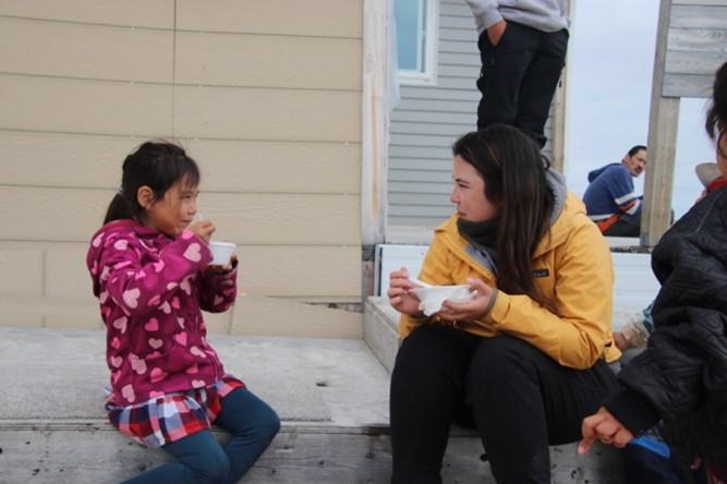 Dënë Cheecham-Uhrich (à droite) discute avec un enfant (à gauche) de la communauté de Cambridge Bay sur l'île Victoria, au Nunavut, alors que tous deux mangent de la soupe et sont assis sur le perron d'un camp de pêche. Crédit photo, Helen Blewett - Coordinatrice des aînés d'Iqaluktuuttiaq. 