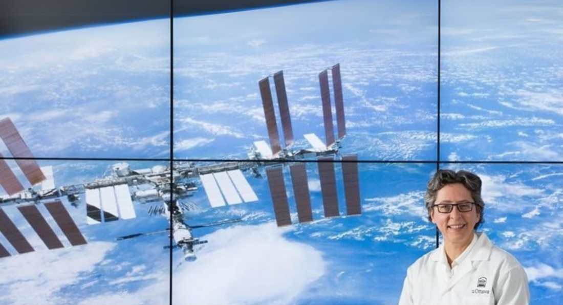 Odette Laneuville, vêtue d'une blouse, debout devant un écran montrant la Station spatiale internationale