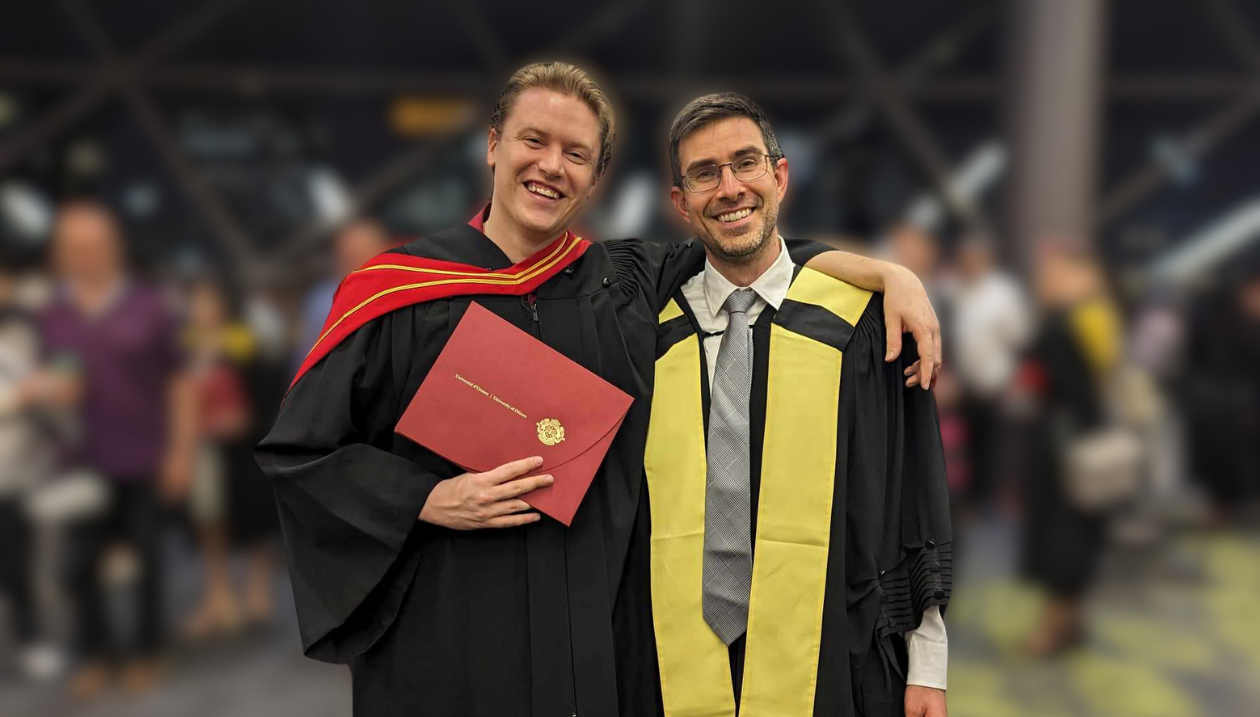 Adam Cook vêtu d'une toge noire avec une capuche rouge et dorée, tenant un diplôme. À sa gauche se tient le professeur Stephen Newman, vêtu d'une toge noire avec une étole jaune. Ils sont entourés d'autres personnes célébrant la remise des diplômes. 