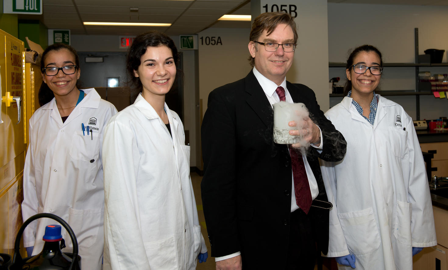 Le professeur Robert Pezacki se tient dans un laboratoire, tenant un bécher rempli de glace carbonique qui dégage de la vapeur visible. Il est entouré de trois doctorantes souriantes en blouse blanche : Noreen Ahmed (à l'extrême gauche), Roxana Filip (à gauche de Pezacki) et Nadine Ahmed (à l'extrême droite). On aperçoit du matériel de laboratoire et des panneaux indicateurs à l'arrière-plan.
