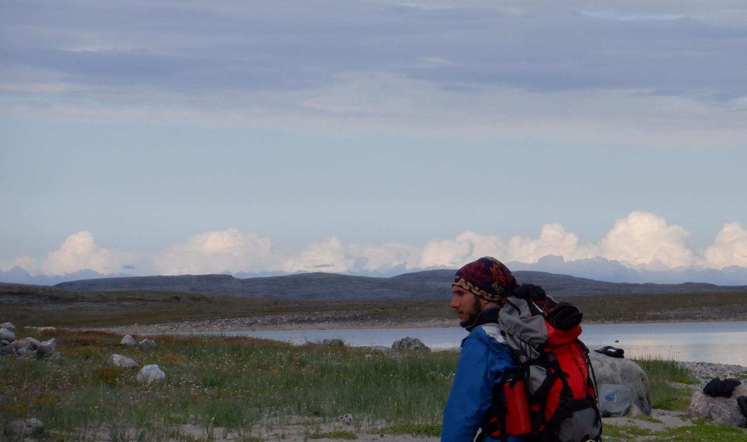Le doctorant Victor Garcia, en expédition sur le terrain, se tient avec un gros sac à dos de randonnée à côté d’un lac arctique isolé près d’Inukjuak, dans le nord du Québec, avec la baie d’Hudson en arrière-plan. 