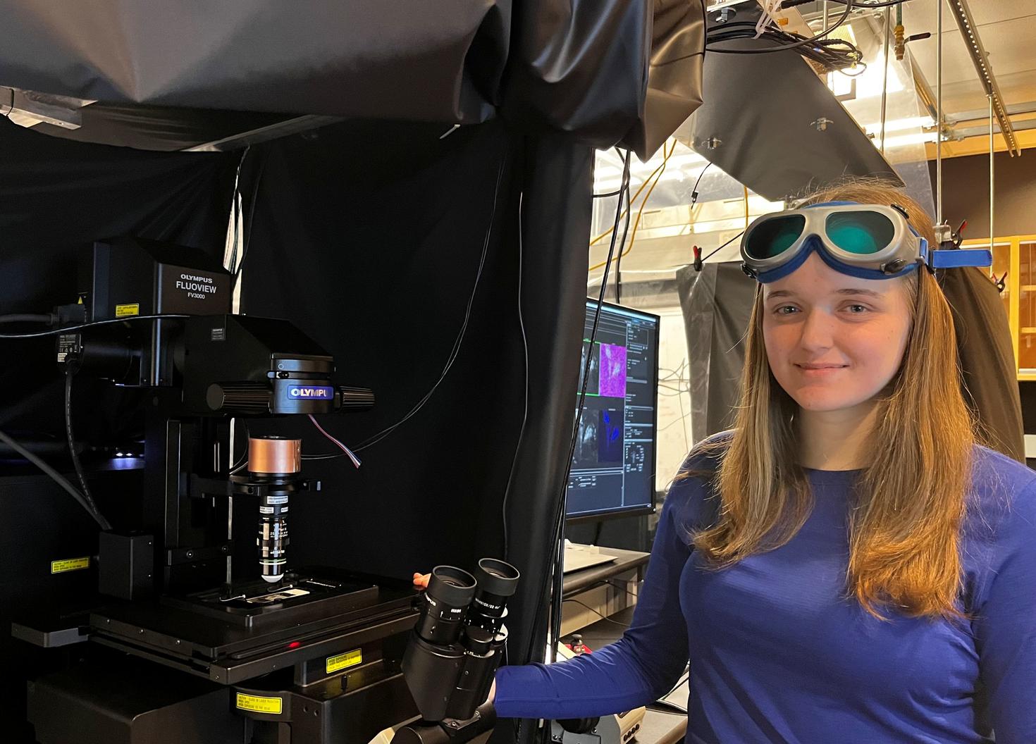 Leah Frackleton se tient à côté d'un microscope à balayage laser dans son laboratoire. Elle porte des lunettes de protection sur le front et tient un microscope dans une main.
