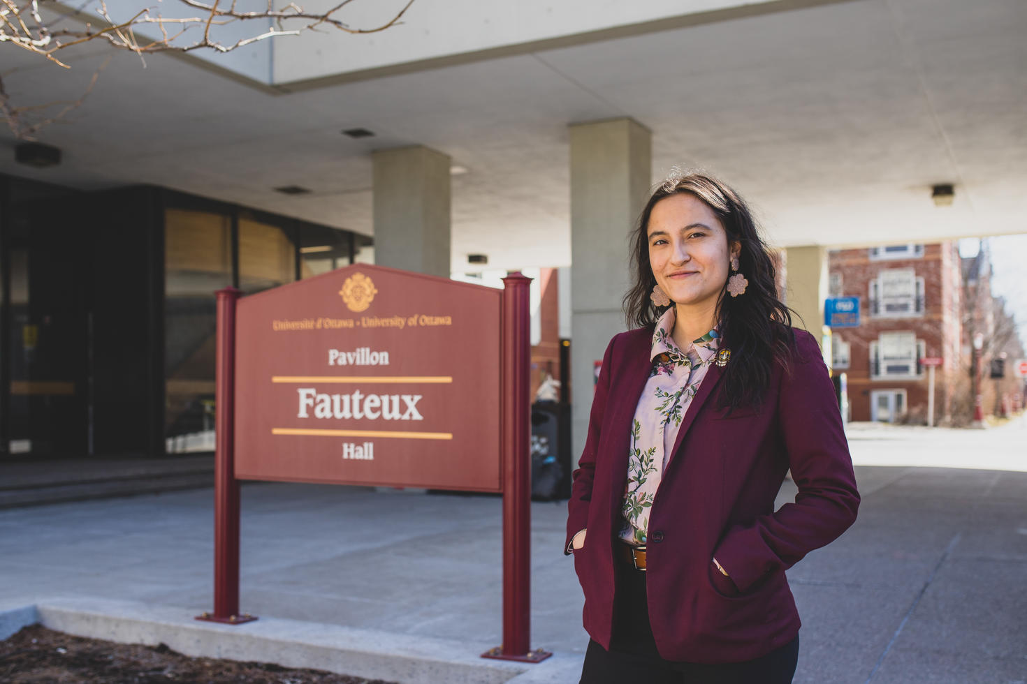 Indigenous student in a purple blazer and flower earrings, smiling and standing in front of and beside the sign for Fauteux Hall, the law building.