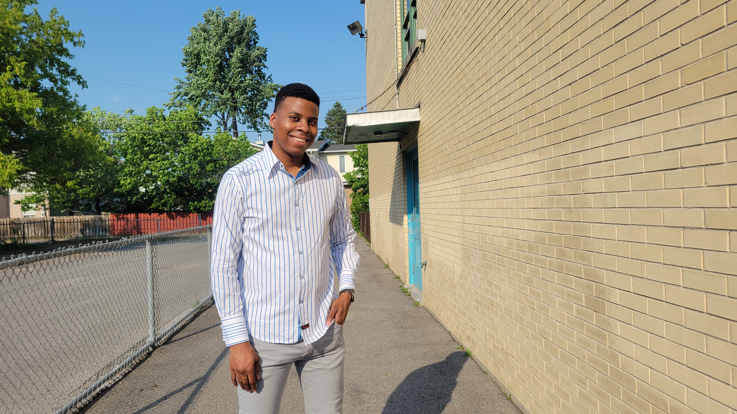 A smiling man standing outside close to a building. Sunny day. Background of trees.