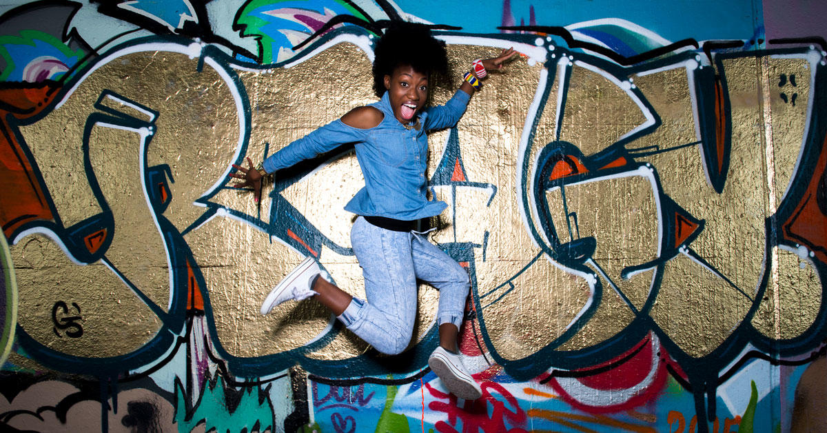 Pearline Barrett-Fraser is jumping and dancing in front of a wall covered with colourful grafifitis.