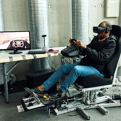 Person sitting in chair with virtual reality goggles on steering a steering wheel.