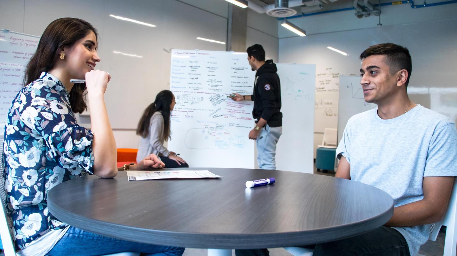 Two people sitting around a table chatting with two others in the background, writing on a whiteboard.