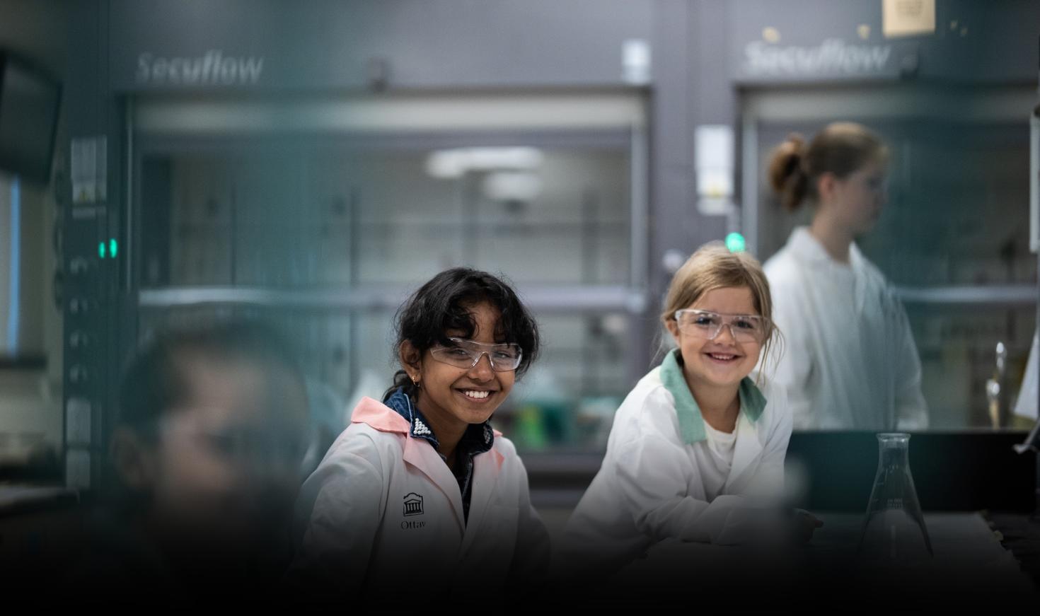 Two young kids with safety glasses and gloves sitting at table doing science experiment with teacher in background.