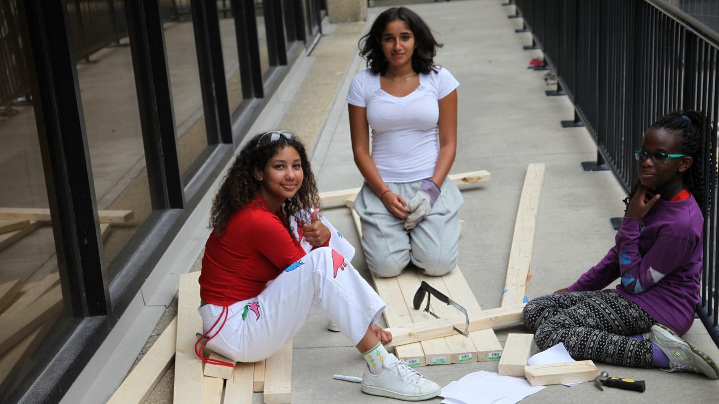 Three girls are working together on a wood construction project. They are sitting on the ground surrounded by wooden planks, tools, and paper.