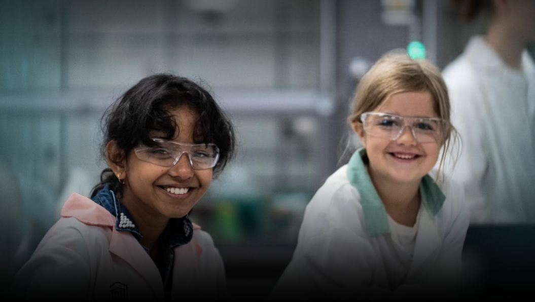 Two young kids with safety glasses and gloves sitting at table doing science experiment with teacher in background.