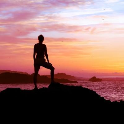 A woman stands on a cliff looking at a sunset