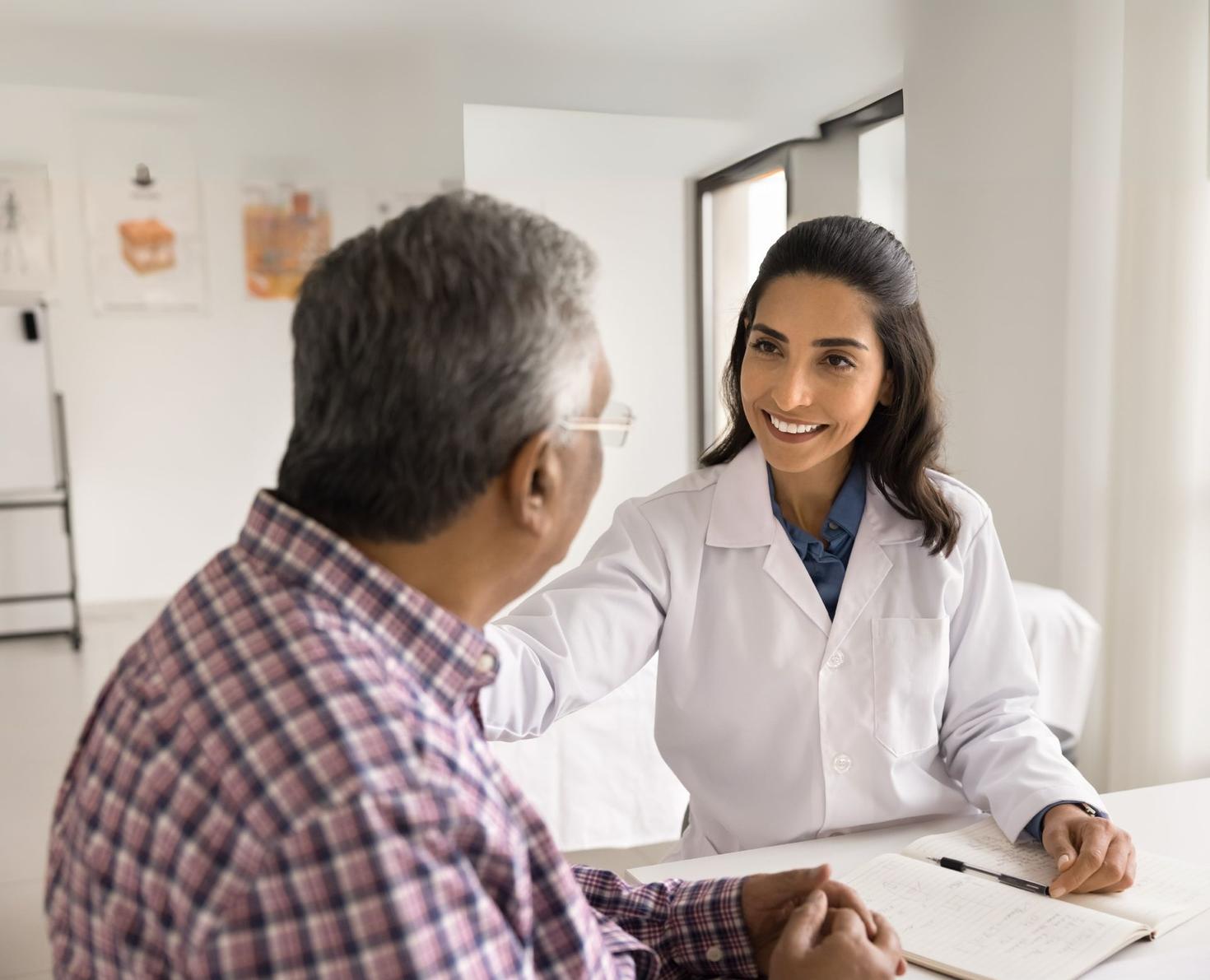 Female health professional in a white coat smiling and speaking with an older man in a plaid shirt during a consultation.