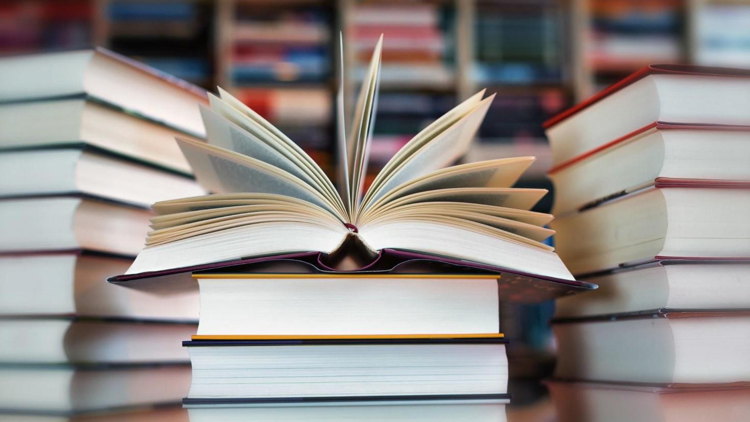 An open book lies on top of a stack of closed books, with more stacked books blurred in the background.