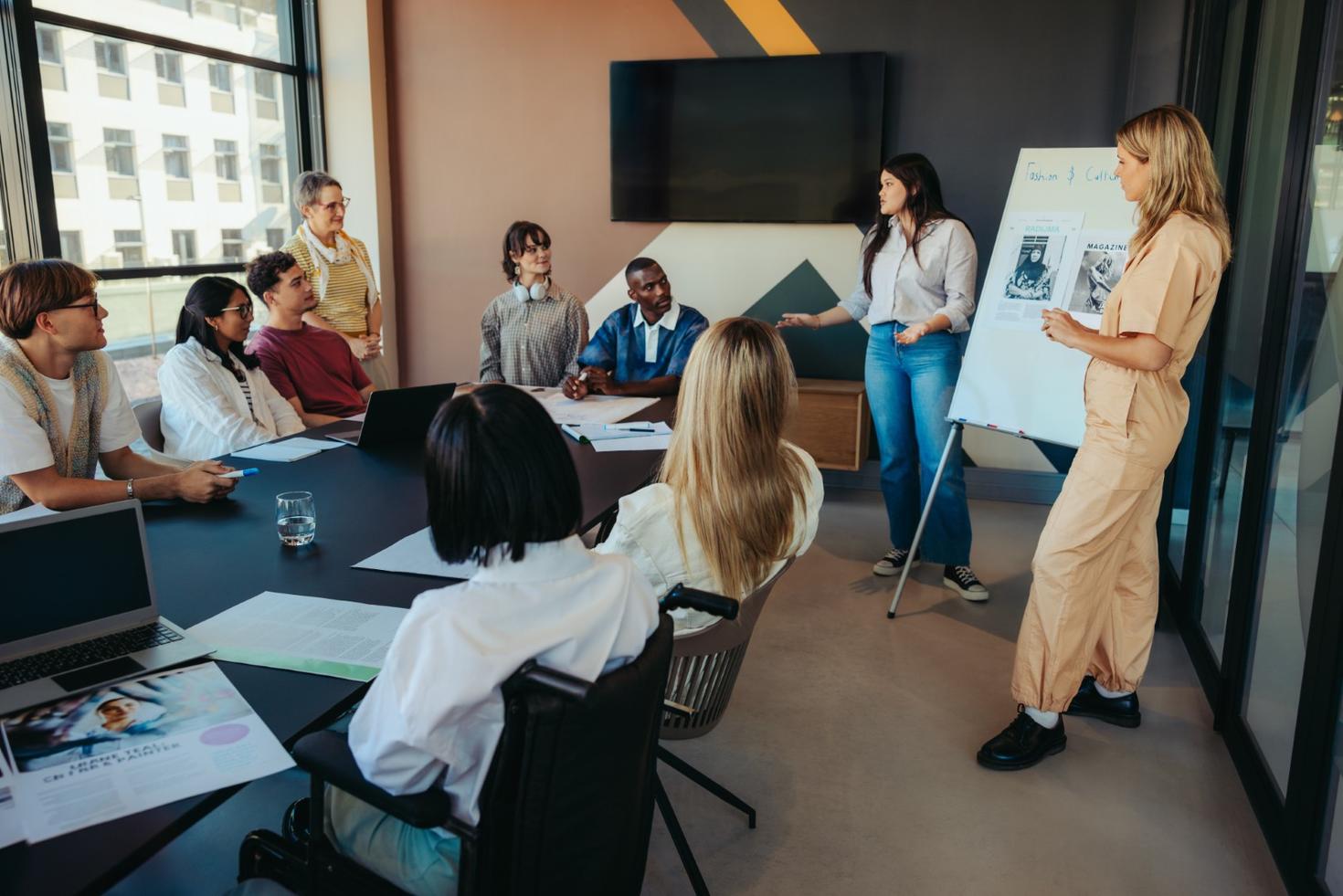 Two women present using a flip chart in a boardroom with several people watching