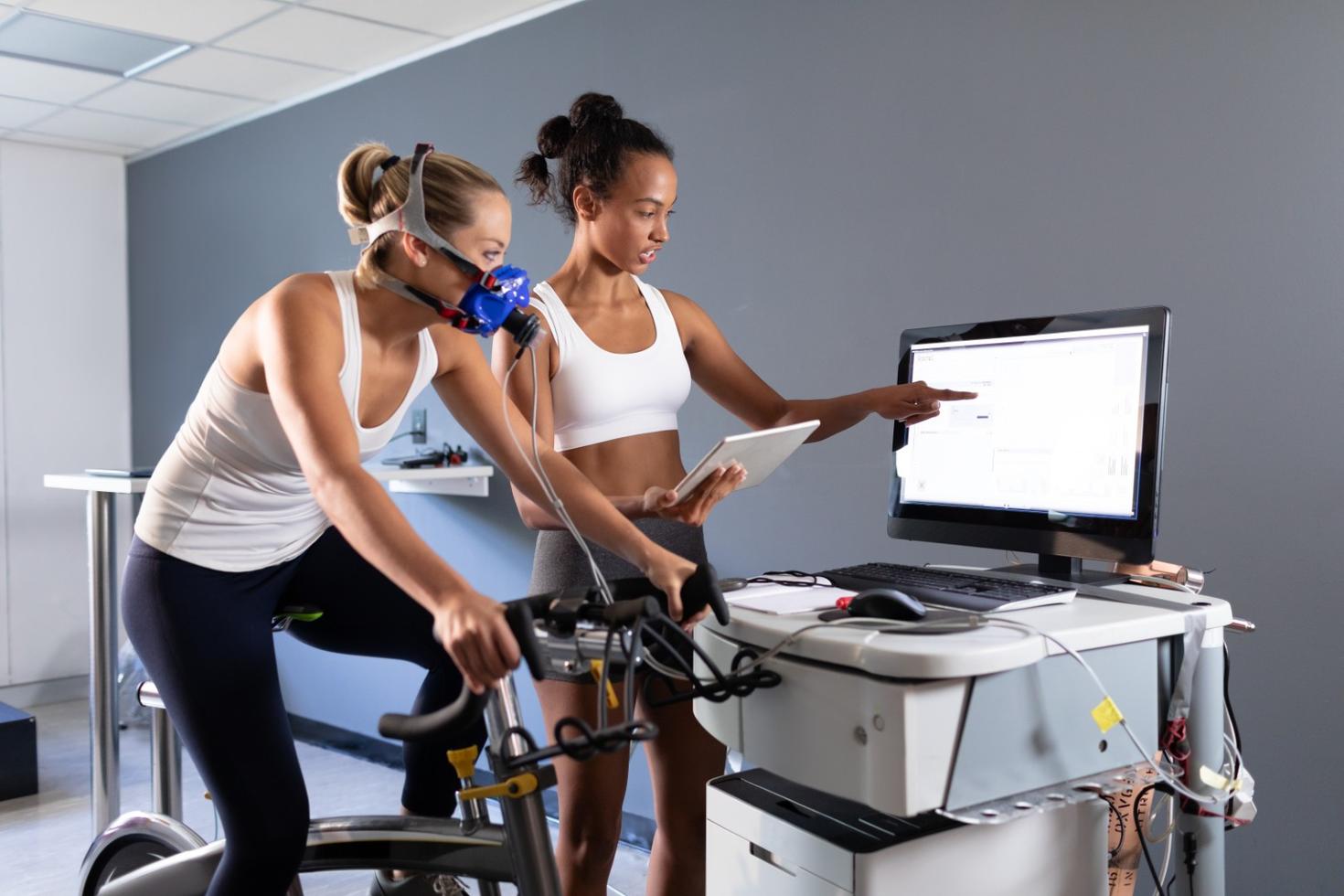 A woman is on a stationary bike with sensors on her and another woman analyzes results on a screen