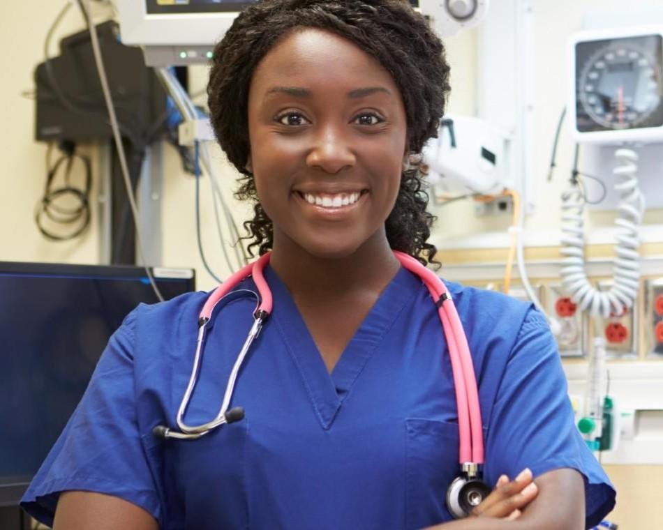 Smiling nurse or doctor in blue scrubs with a stethoscope around her neck, standing in a medical setting.