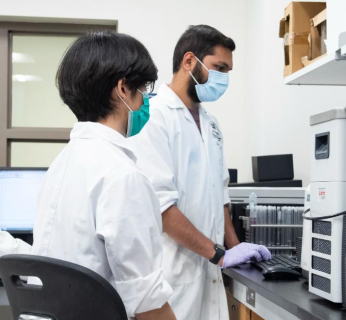 Two researchers wearing lab coats and masks working with lab equipment and a computer in a research lab.