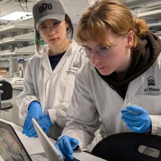 Two students in lab coats and gloves, looking at a laptop in a lab.