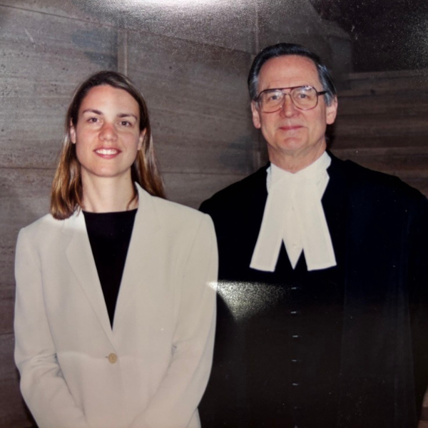 A man and a woman stand facing the camera. The woman is wearing a suit, the man is wearing judge's robes.