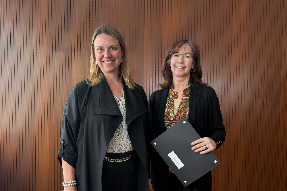 Two women stand, facing forward and smiling. One is holding a folder.