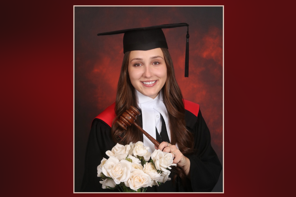 A young woman, wearing a black robe and black graduation cap, holds a bouquet of white roses.