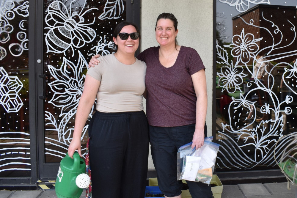 Two women stand outside, in front of a glass wall that is decorated with drawings of plants, flowers and insects. One woman is holding a watering can and wearing sunglasses. The other woman is holding a bag full of seed packets.