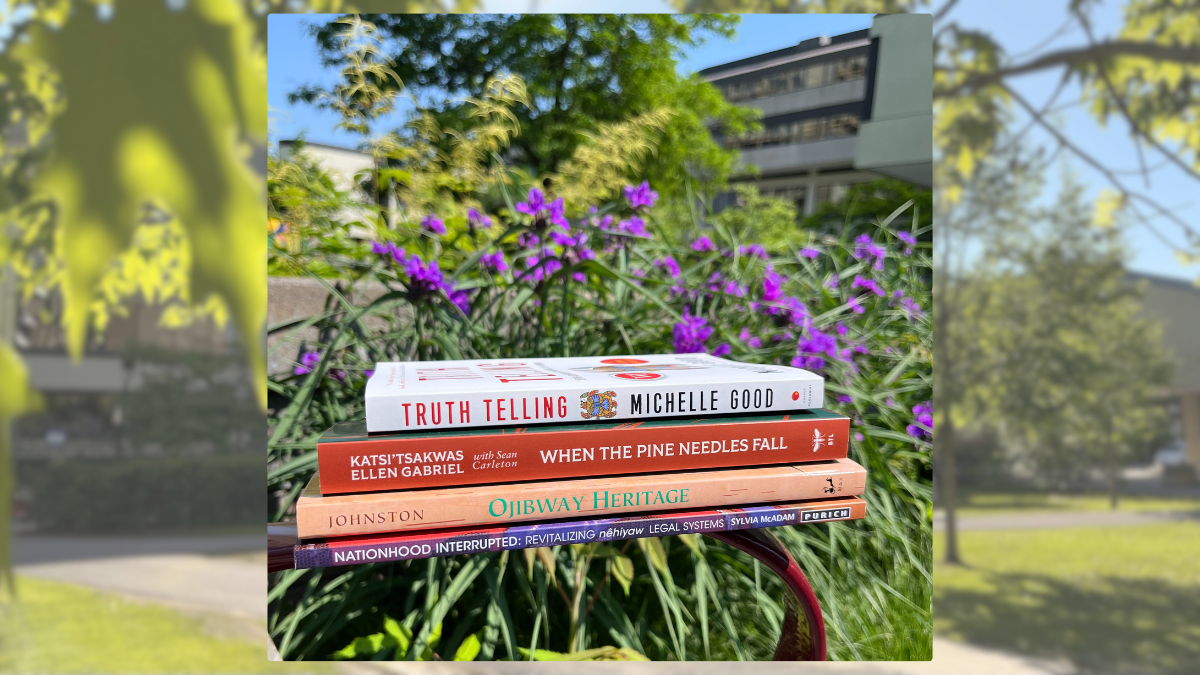 A stack of books sits on the edge of a bench, with grass and flowers in the background