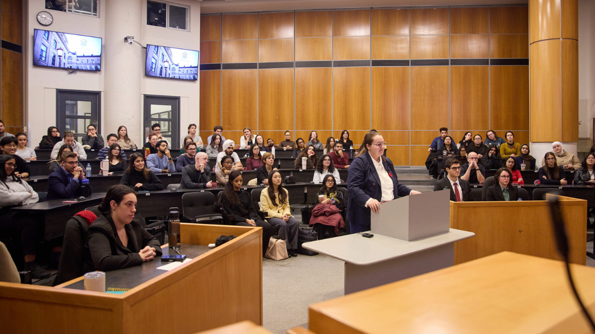 A classroom full of university students, one young woman stands at the front, at a podium