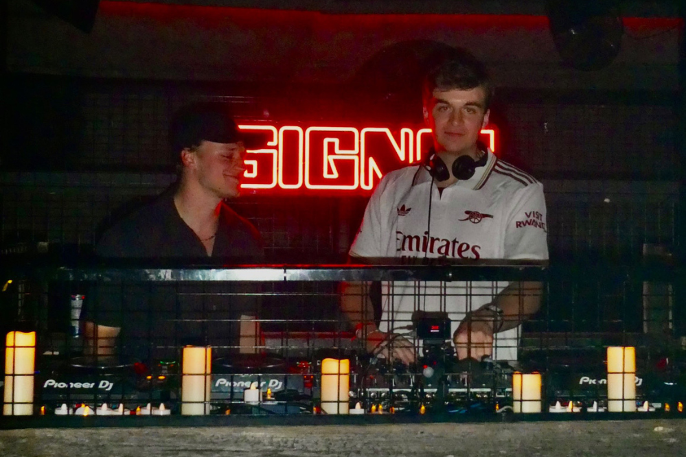 Two young men stand behind a DJ table. One of them is smiling at the camera.