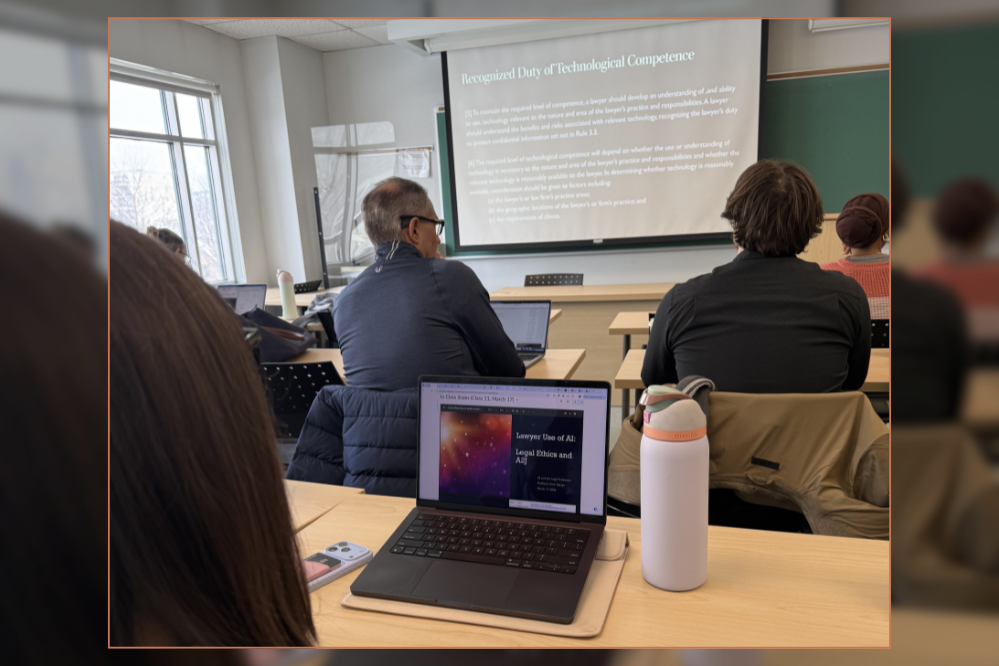 View from the back of a classroom. Students are looking at a large screen and many have laptops on the table in front of them.