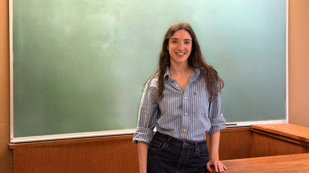 A young woman stands in front of a blackboard, leaning against a desk