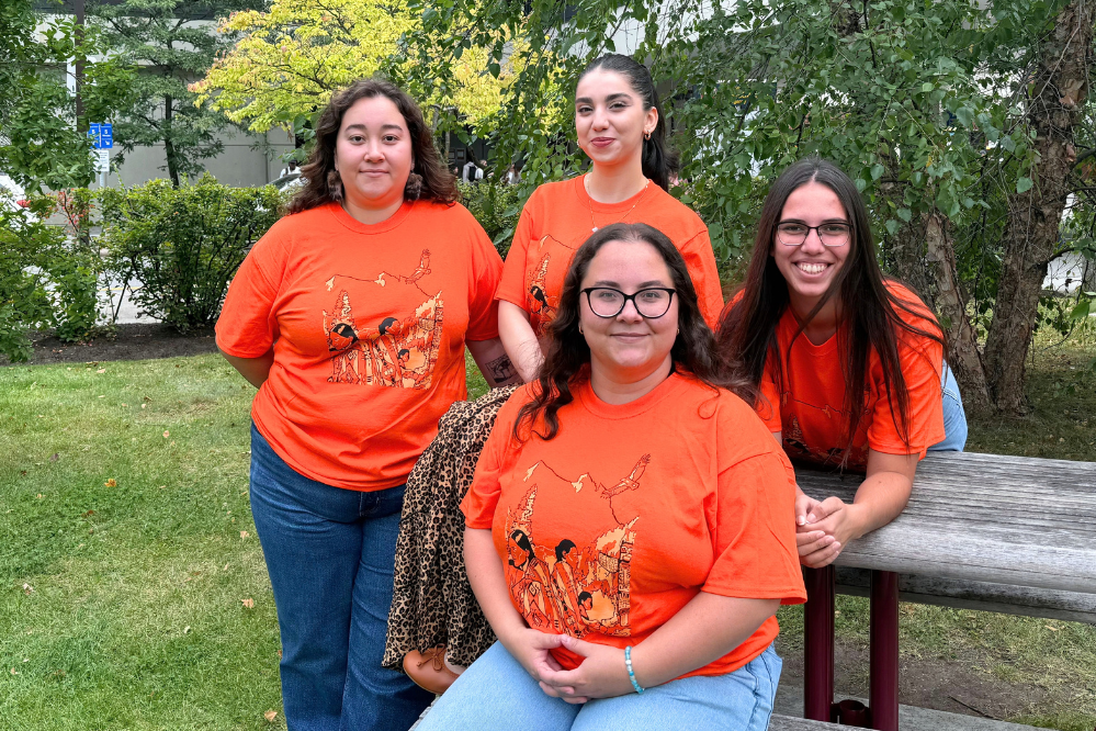 Four young women, all wearing the same Orange Tshirt design, sit and stand near a picnic table outside.