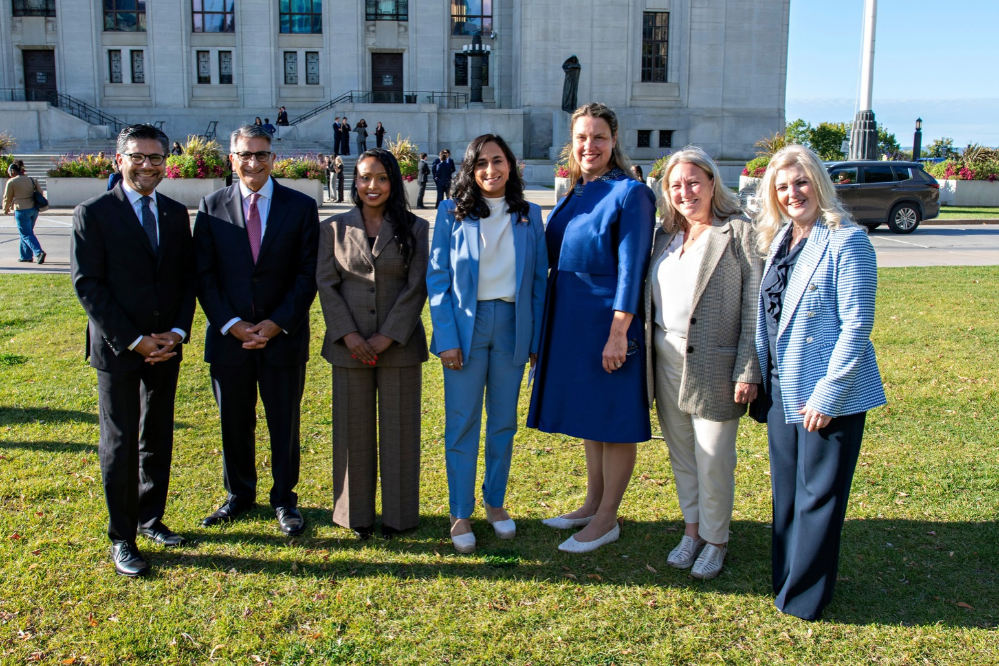 Many people in business attire stand facing the camera. They are outside and it is a sunny day. The people are smiling.